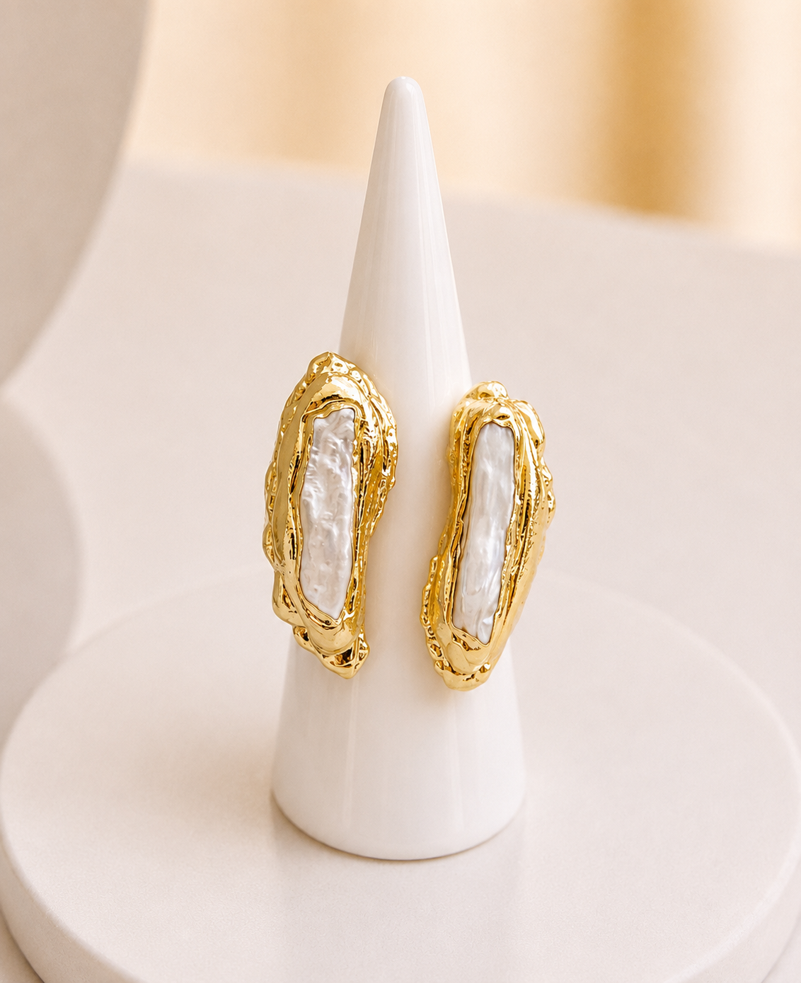 A gold-toned statement ring with irregular organic borders and central white mother-of-pearl inlays, displayed on a white cone stand against a soft beige background.