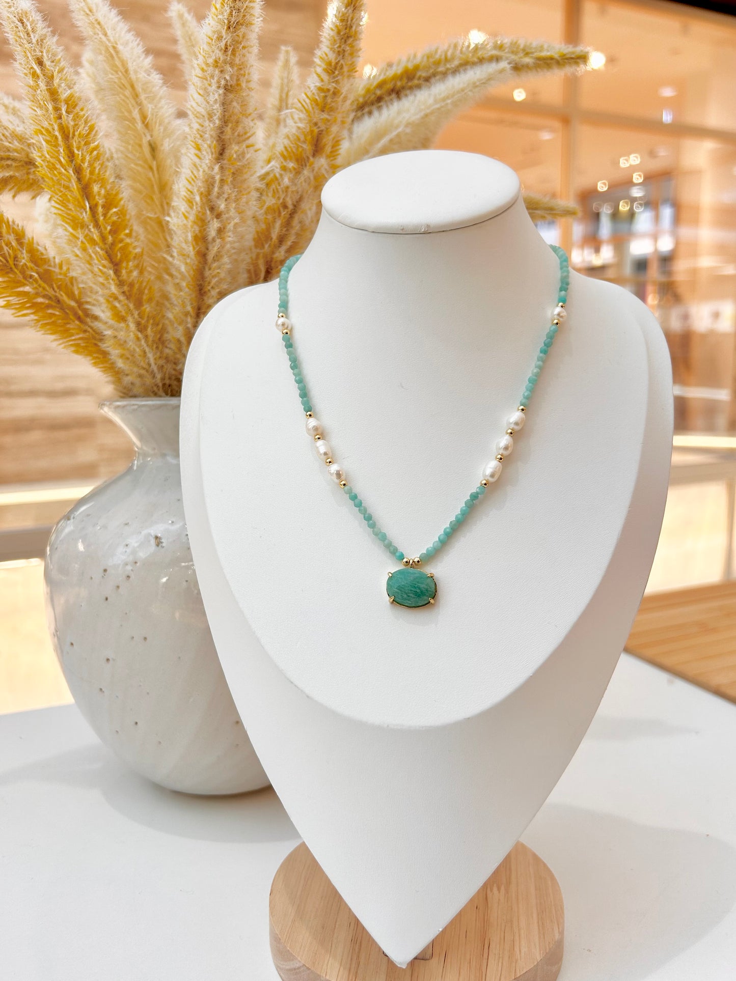 
                  
                    A beaded necklace with amazonite turquoise-green beads, small white pearl-like beads, and a central oval turquoise pendant, displayed on a white jewellery bust. The bust is set on a light wood base with dried pampas grass and a textured white vase in the softly blurred background.
                  
                