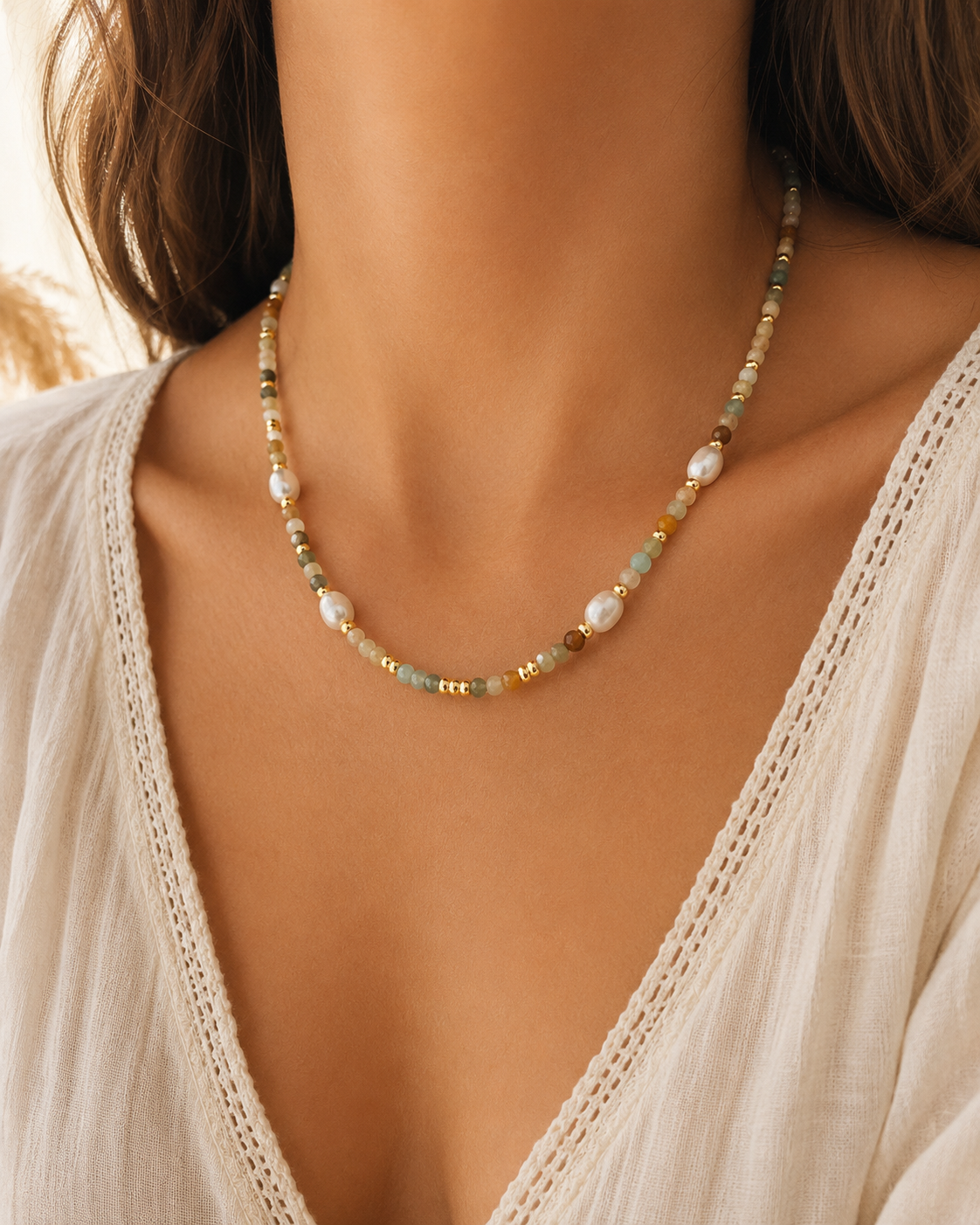 A close-up of a woman wearing a delicate beaded necklace with white pearls and small mixed amazonite stones paired with a cream-colored lace-trimmed top.