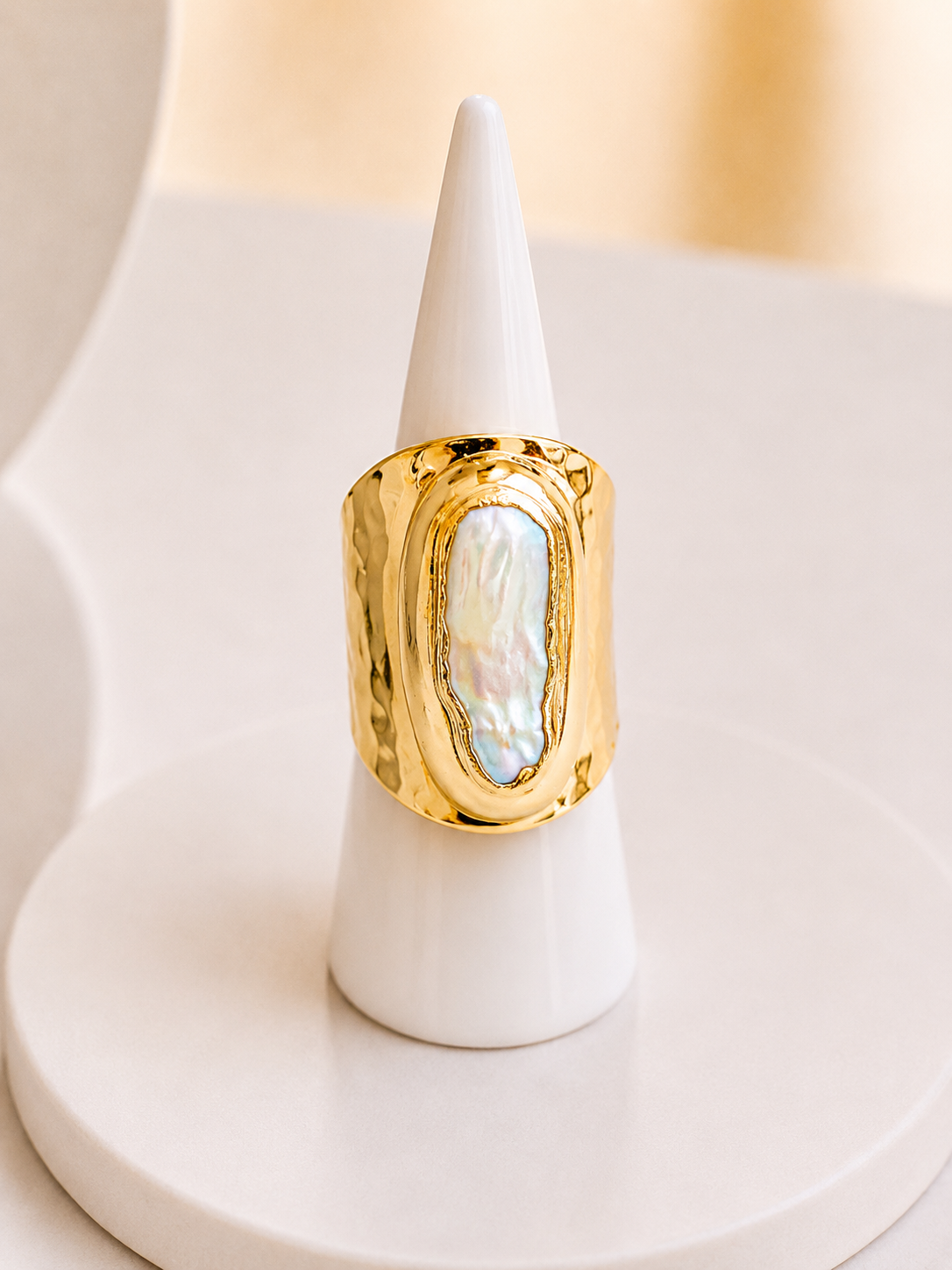A chunky gold ring with a large, irregularly shaped iridescent mother-of-pearl inlay, displayed on a white cone stand.