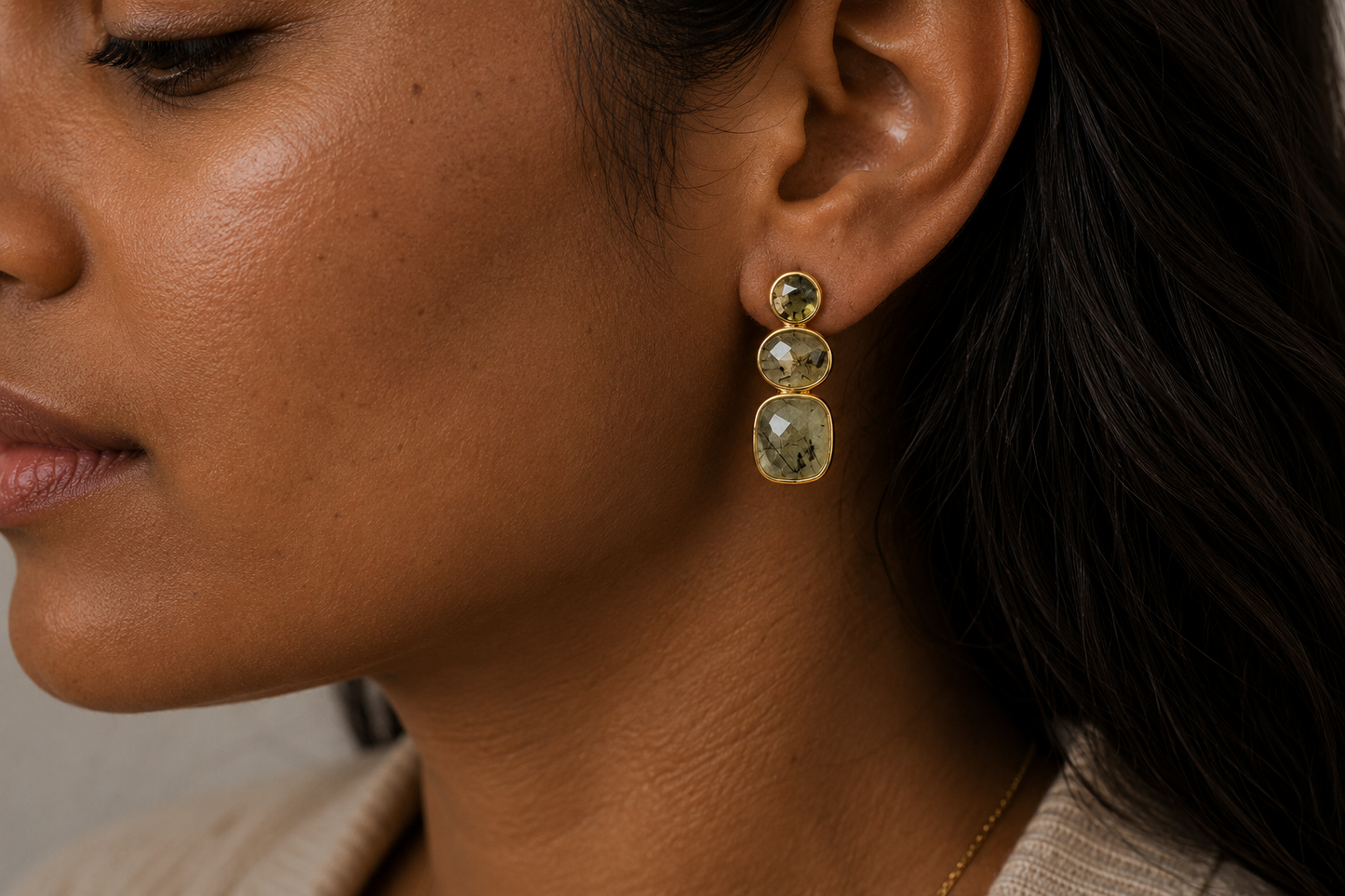 A close-up side profile of a woman wearing a gold drop earring with two stacked oval green gemstones.