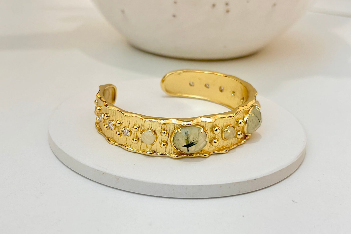 
                  
                    A Gold-plated textured open cuff bracelet featuring natural prehnite gemstones and delicate crystal accents, displayed on a white jewellery stand.
                  
                