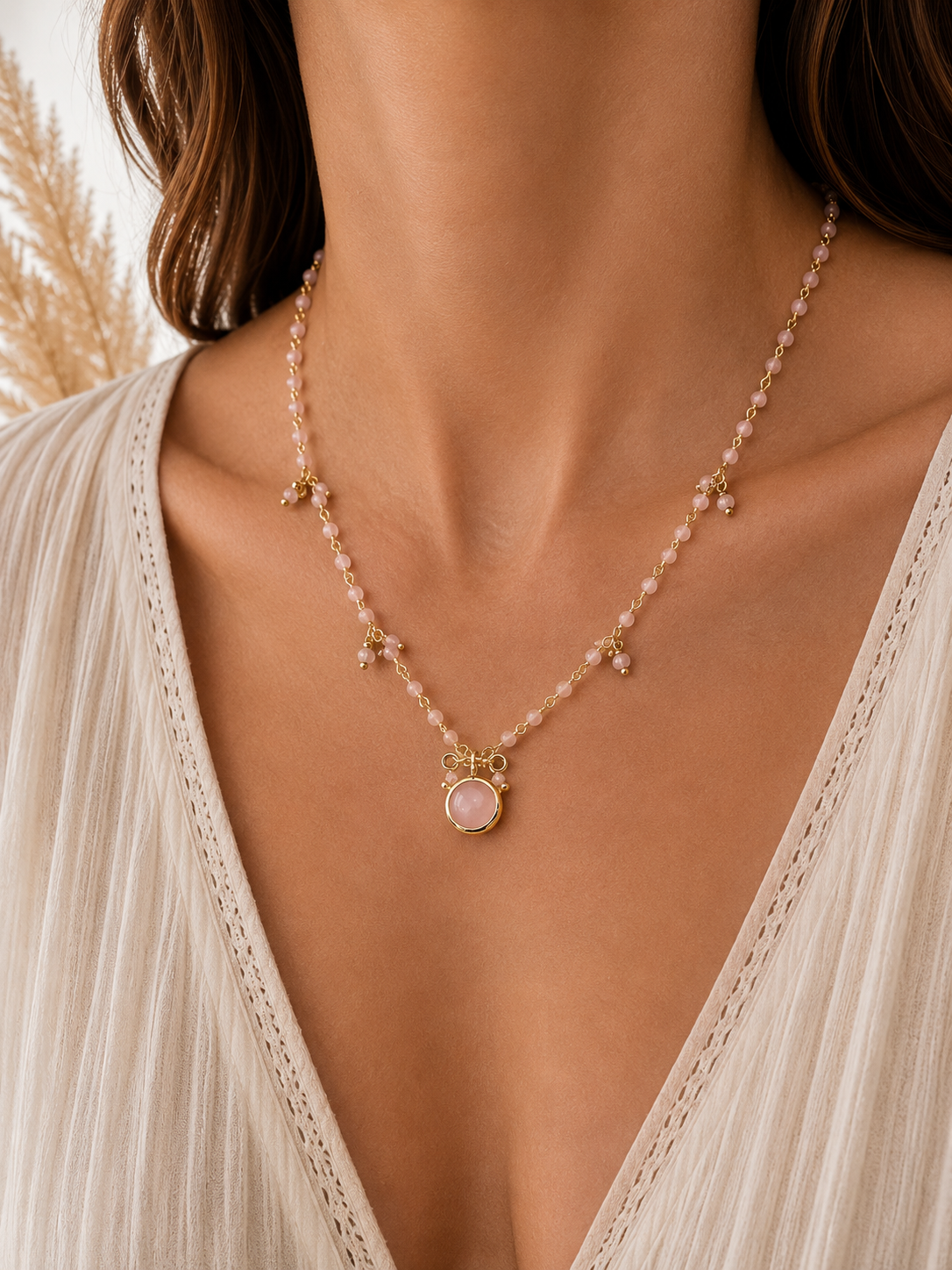 
                  
                    A close-up of a model wearing a delicate gold and pink beaded necklace with a central oval pink rose quartz stone pendant, styled with a cream-colored lace top.
                  
                