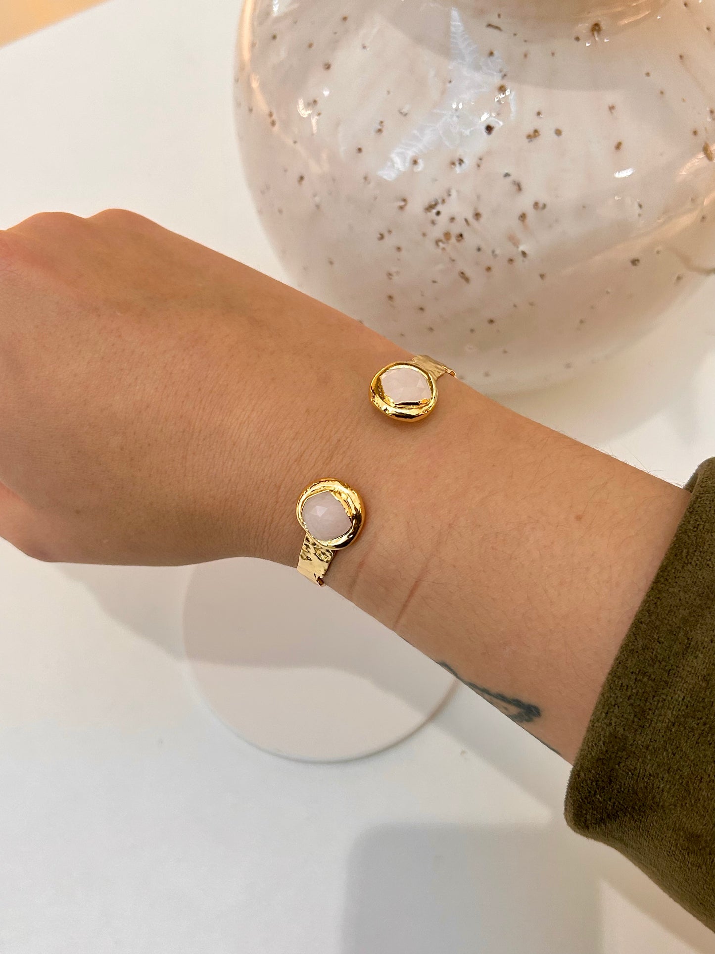 A close-up of a person's wrist wearing gold-toned cuff with oval pale rose quartz stone faces, shown against a light tabletop with a decorative vase in the background.