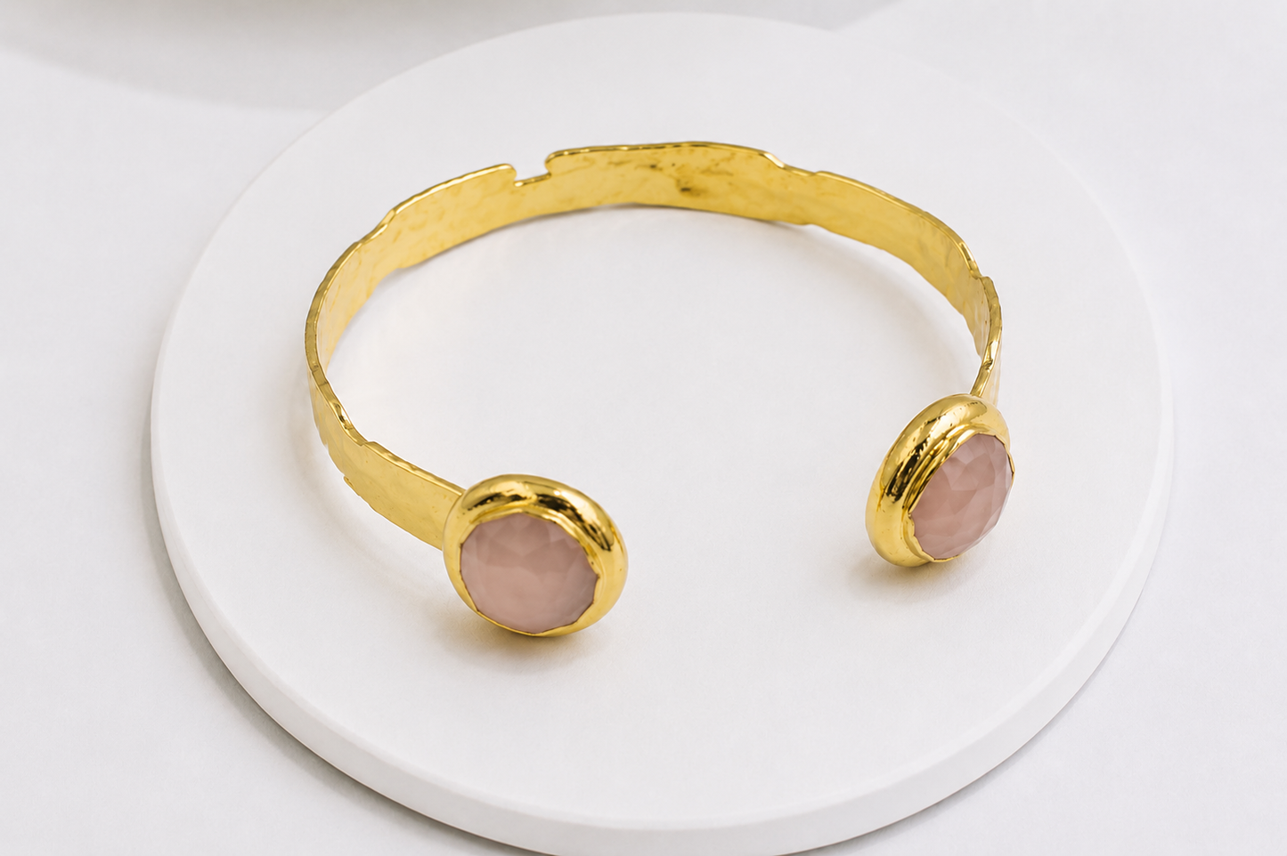 A gold-toned open cuff bracelet with two round pale rose quartz stone accents, displayed on a white round pedestal against a white background.