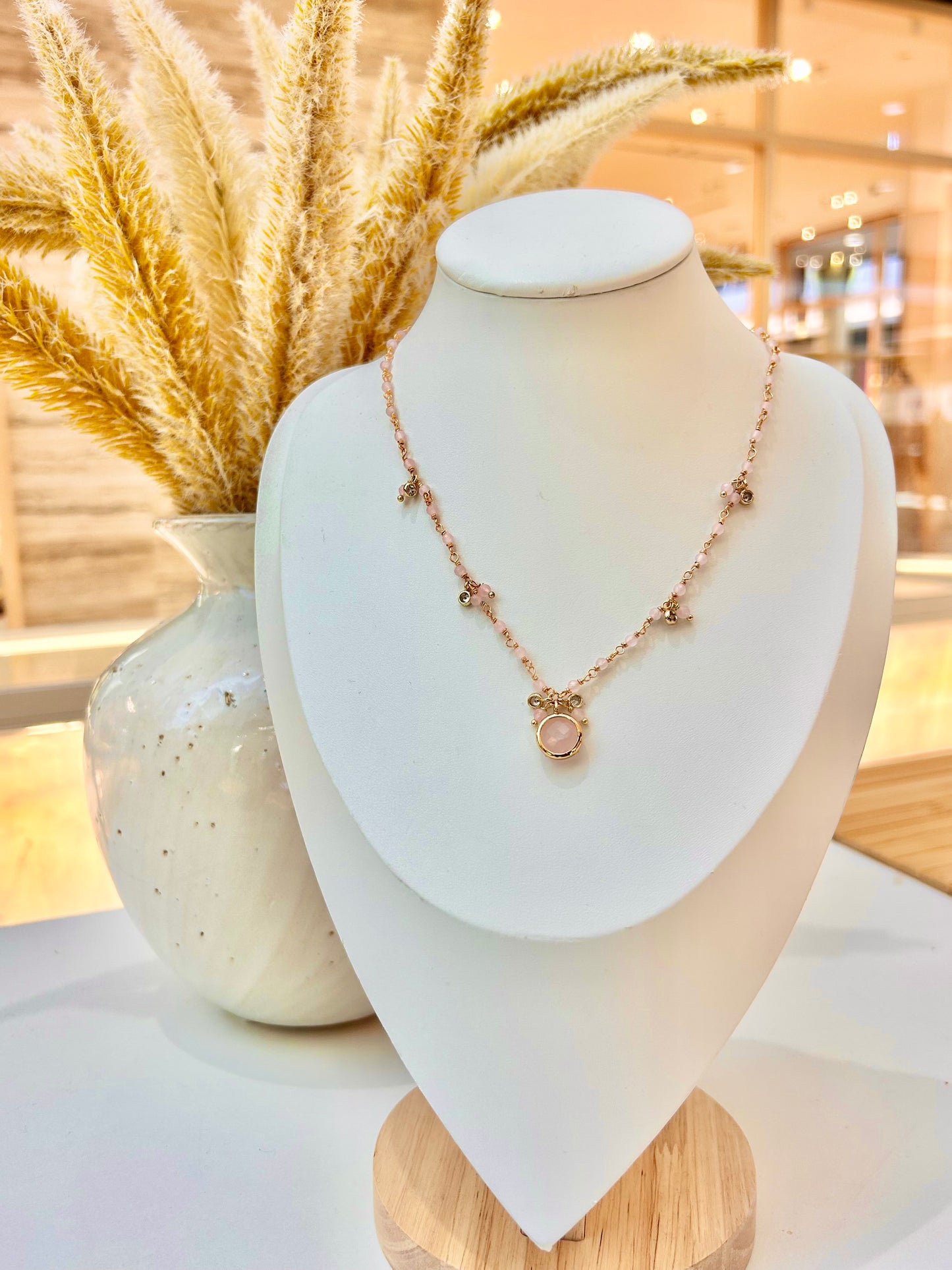 
                  
                    A delicate rose gold necklace with a central circular pendant displayed on a white jewelry bust. The bust is styled with a ceramic vase of dried pampas grass in the background.
                  
                