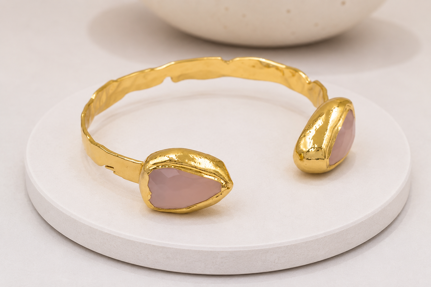A gold cuff bracelet with two teardrop-shaped pale pink rose quartz gemstone inlays, displayed on a round white coaster.