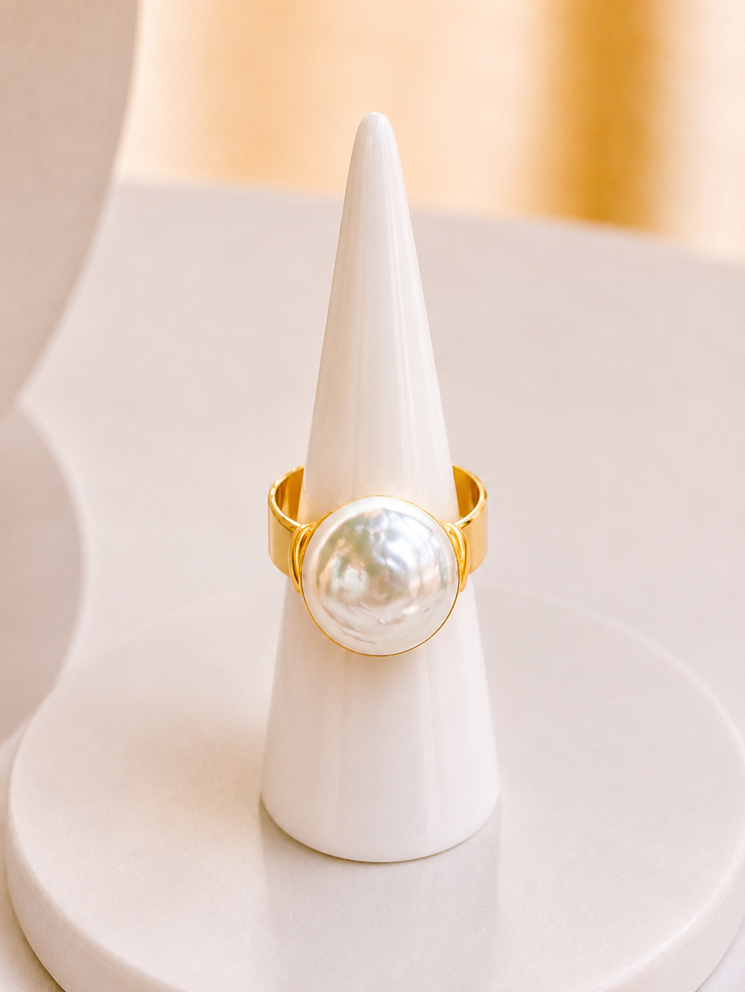 A gold ring with a large, lustrous white pearl bezel-set in the center, displayed on a white cone jewelry stand.