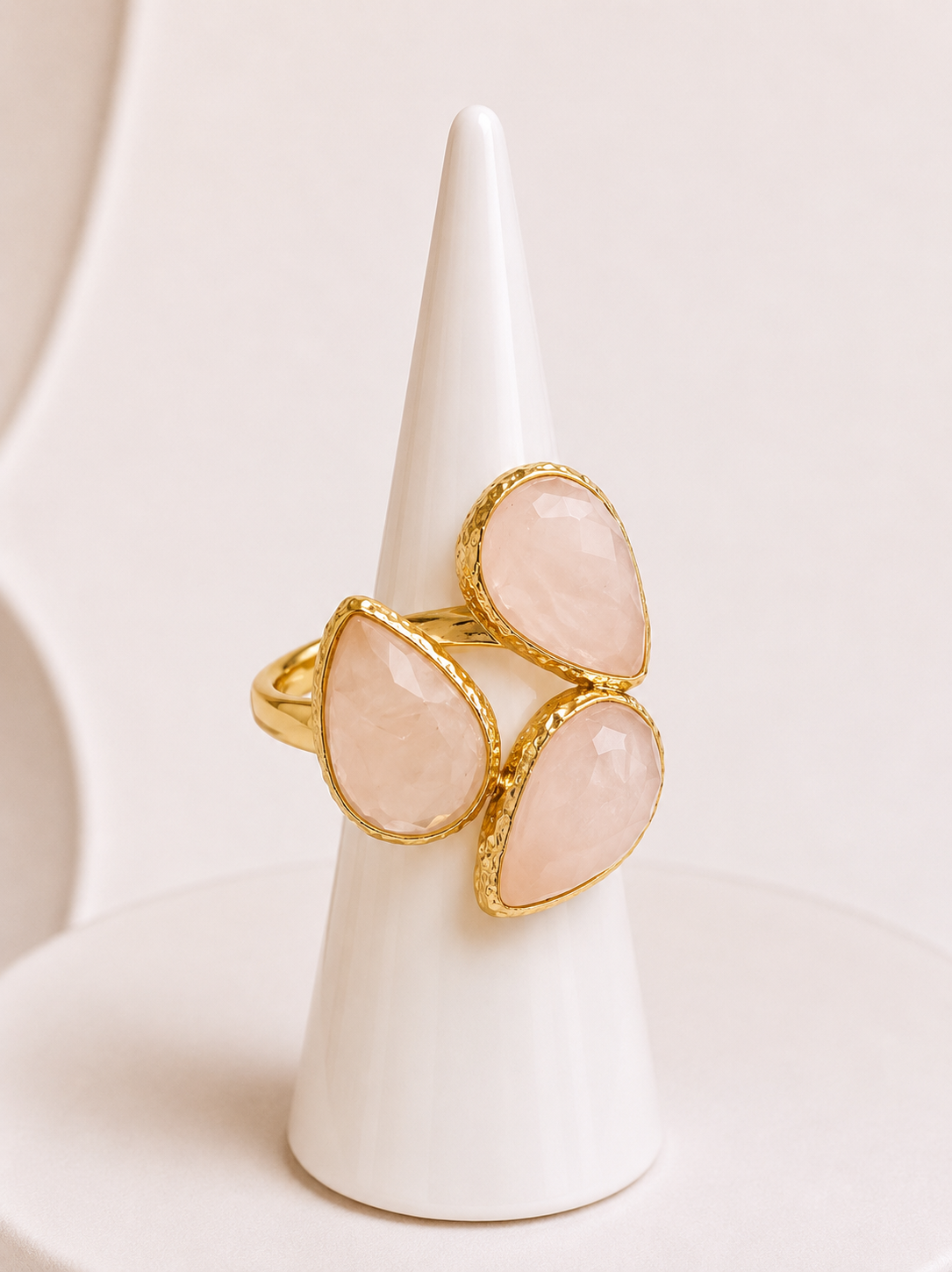 A gold ring with three teardrop-shaped pale pink rose quartz stones, displayed on a white cone stand against a soft cream background.