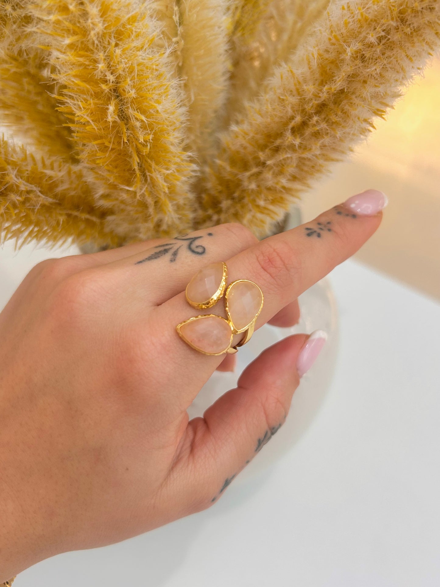 A gold ring with three teardrop-shaped pale pink rose quartz stones worn on a person's finger, shown against a background of dried pampas grass.