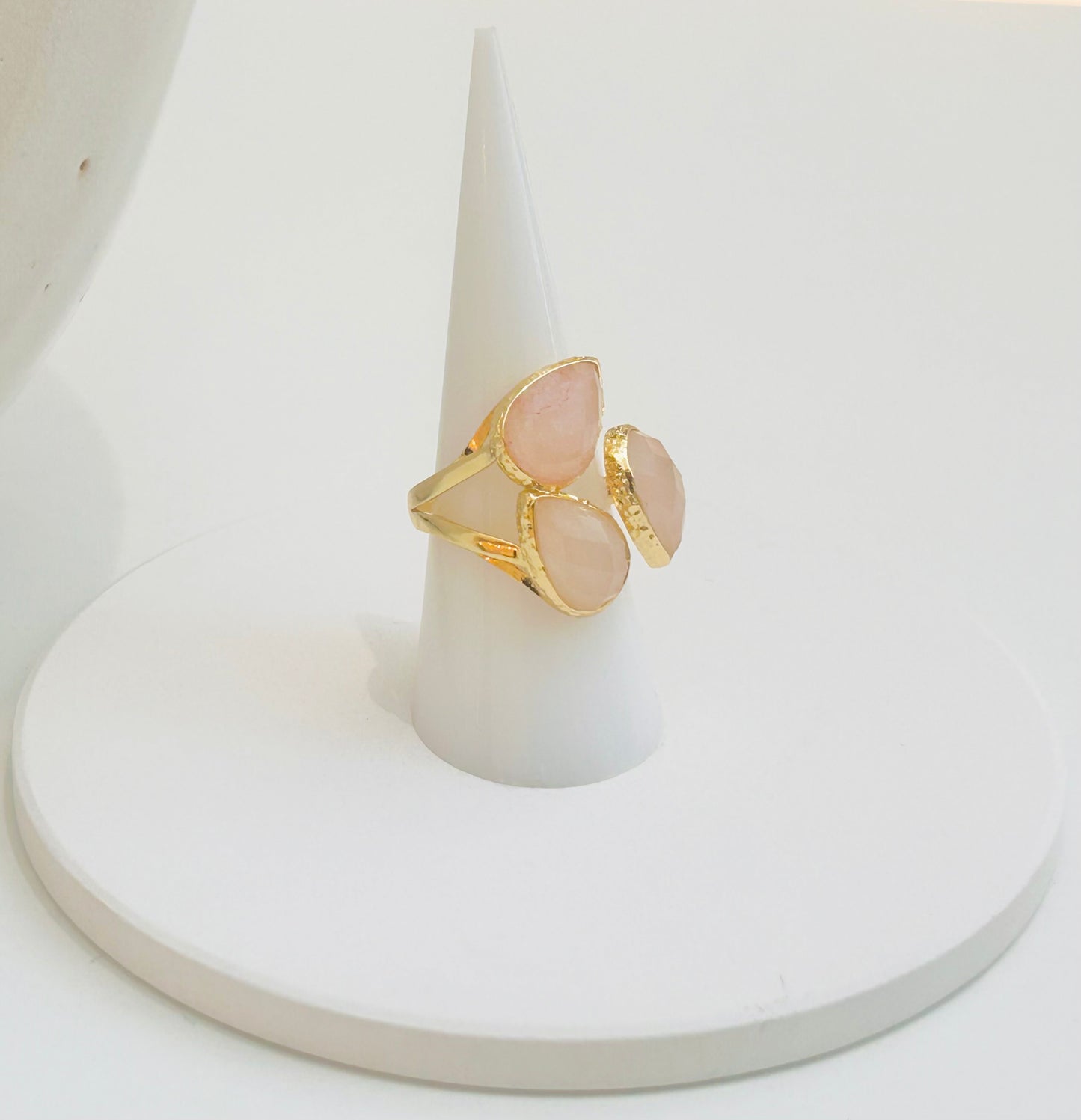 
                  
                    A gold ring with three oval pale pink rose quartz  stones, displayed on a white cone stand against a white background.
                  
                