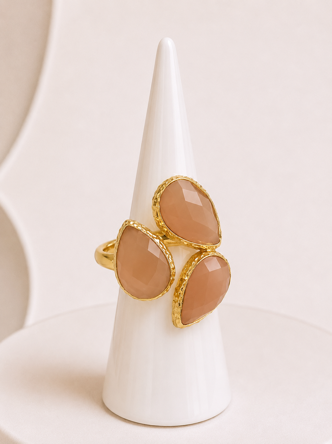 A gold ring with three teardrop-shaped sunstone stones, displayed on a white cone-shaped jewellery stand against a soft cream background.