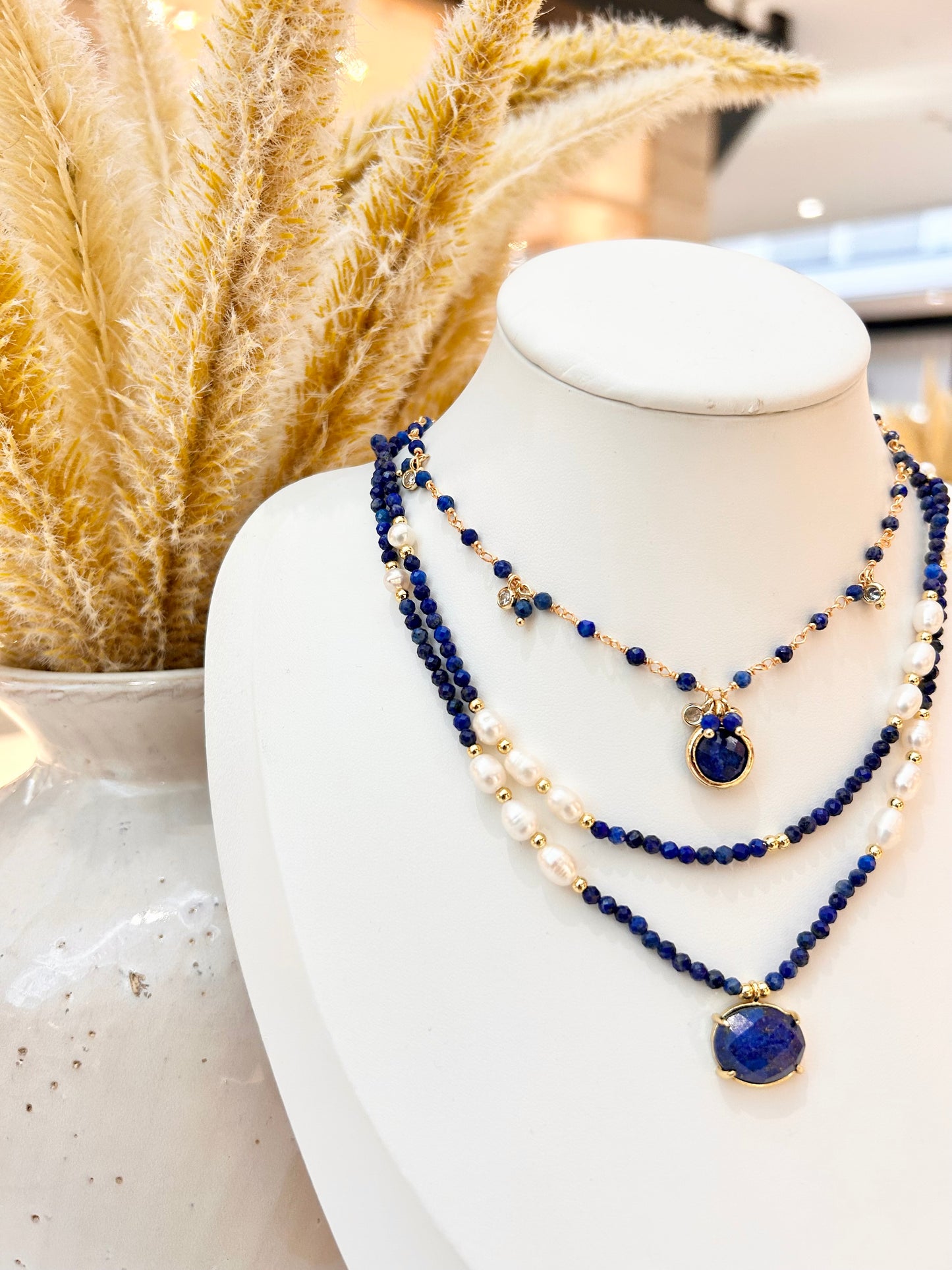 
                  
                    A three-layer beaded necklace set displayed on a white mannequin bust. The necklace features dark blue lapis lazuli stones, white pearls, and small gold-toned spacer beads and pendants.
                  
                