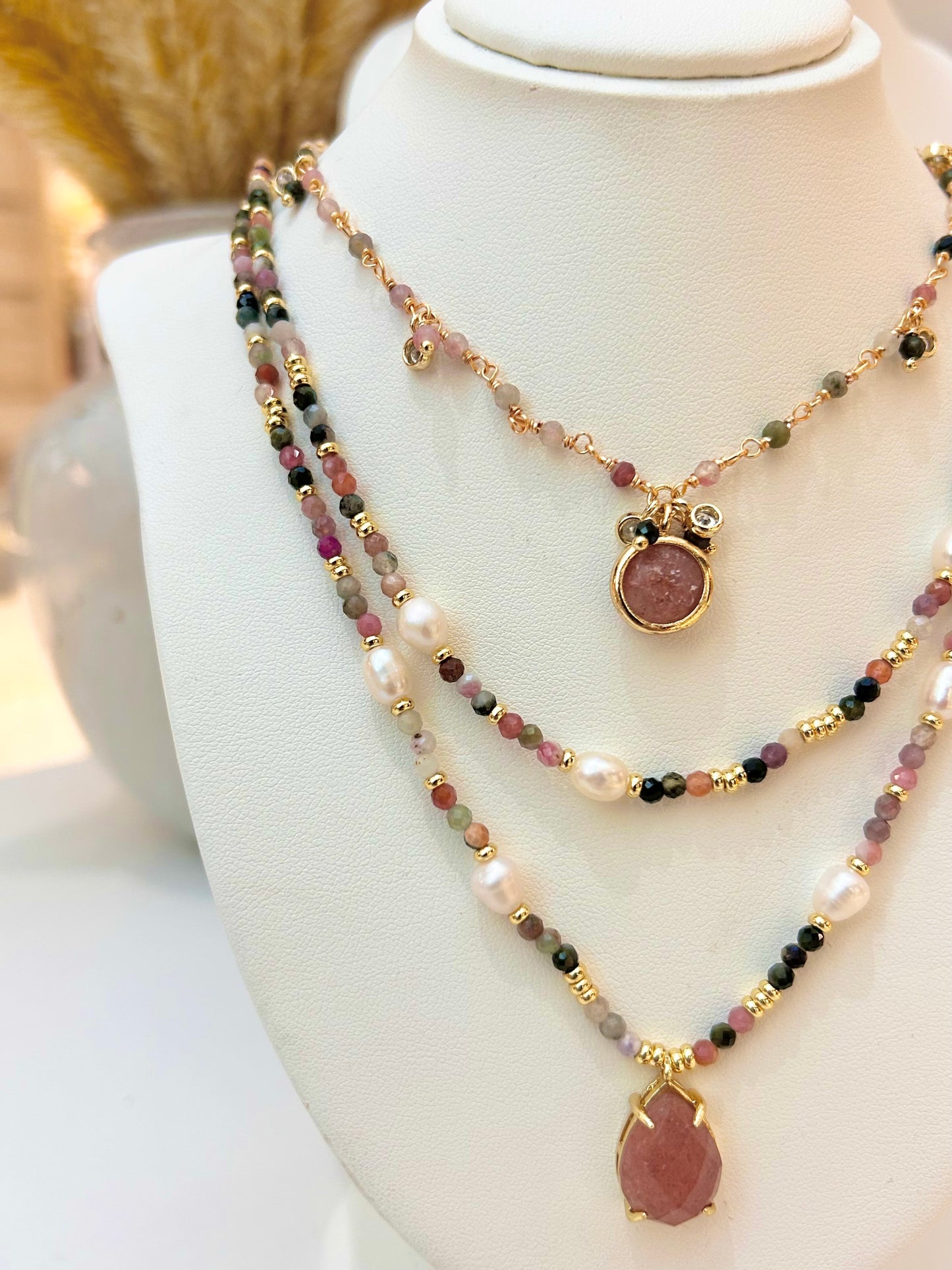 
                  
                    A multi-strand beaded necklace displayed on a white jewellery bust. The necklace features gold-tone chains, white freshwater pearls beads, faceted beads in pink, green, and black, and two teardrop-shaped pink stone pendants in gold-tone settings.
                  
                