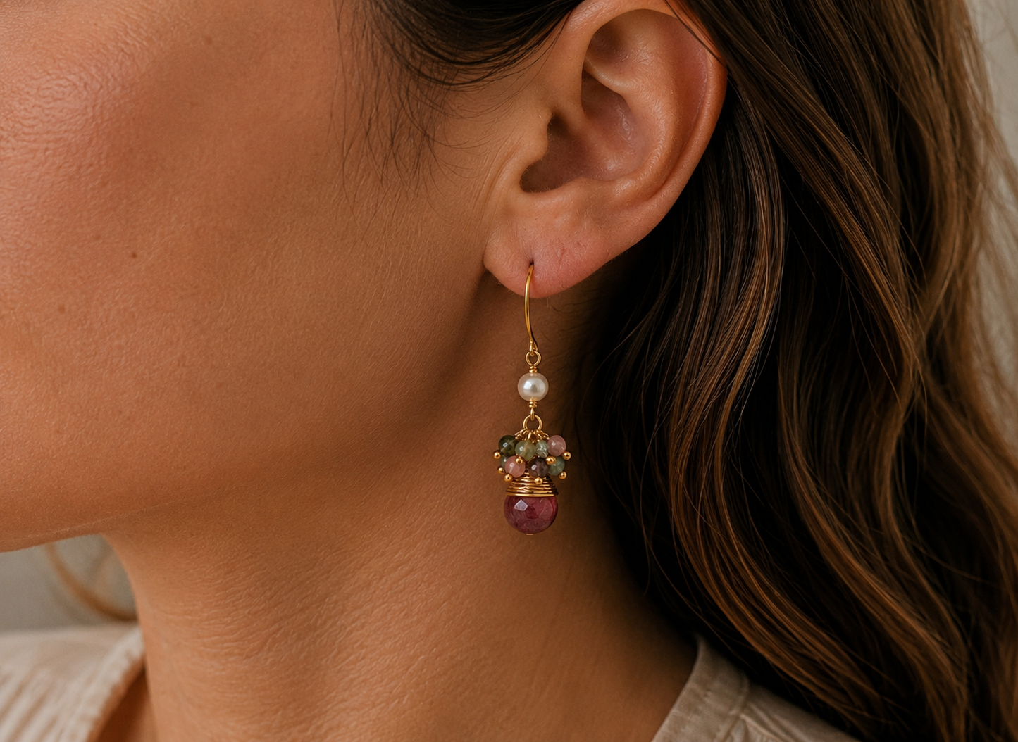 A close-up of a woman's ear wearing a gold dangle earring with a white pearl, small strawberry quartz beads, and a faceted strawberry quartz gemstone.