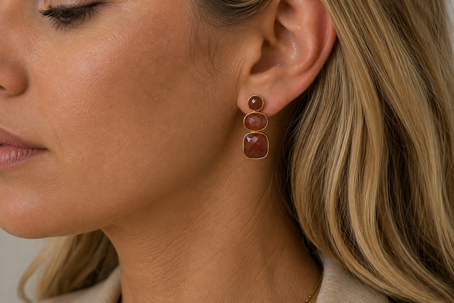 A close-up of a woman's ear wearing a gold drop earring with three stacked oval strawberry quartz gemstones in a warm reddish-brown colour.