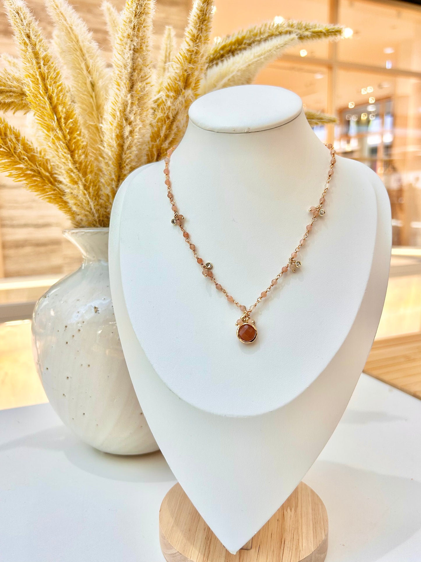 
                  
                    A delicate rose gold necklace with a small round sunstone pendant displayed on a white jewellery  bust. The bust is set on a light wood base, with a white ceramic vase of dried pampas grass in the background.
                  
                