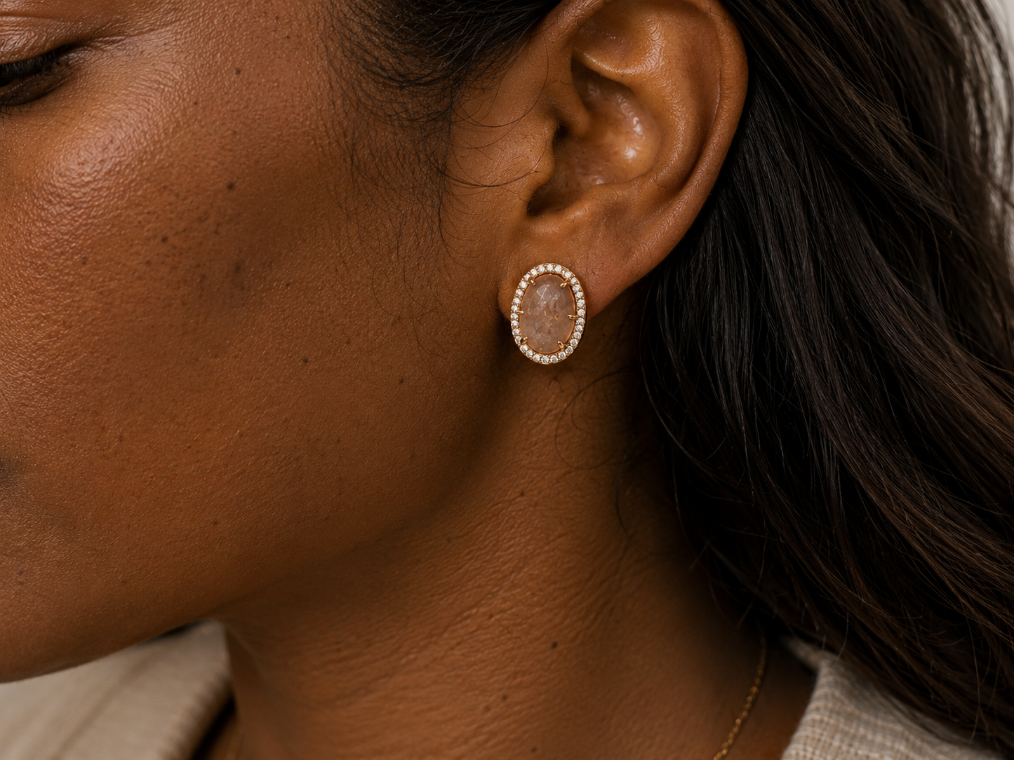 A close-up side profile of a woman wearing a small oval stud earring with a gold-toned border and a sunstone , shown against her ear and dark hair.