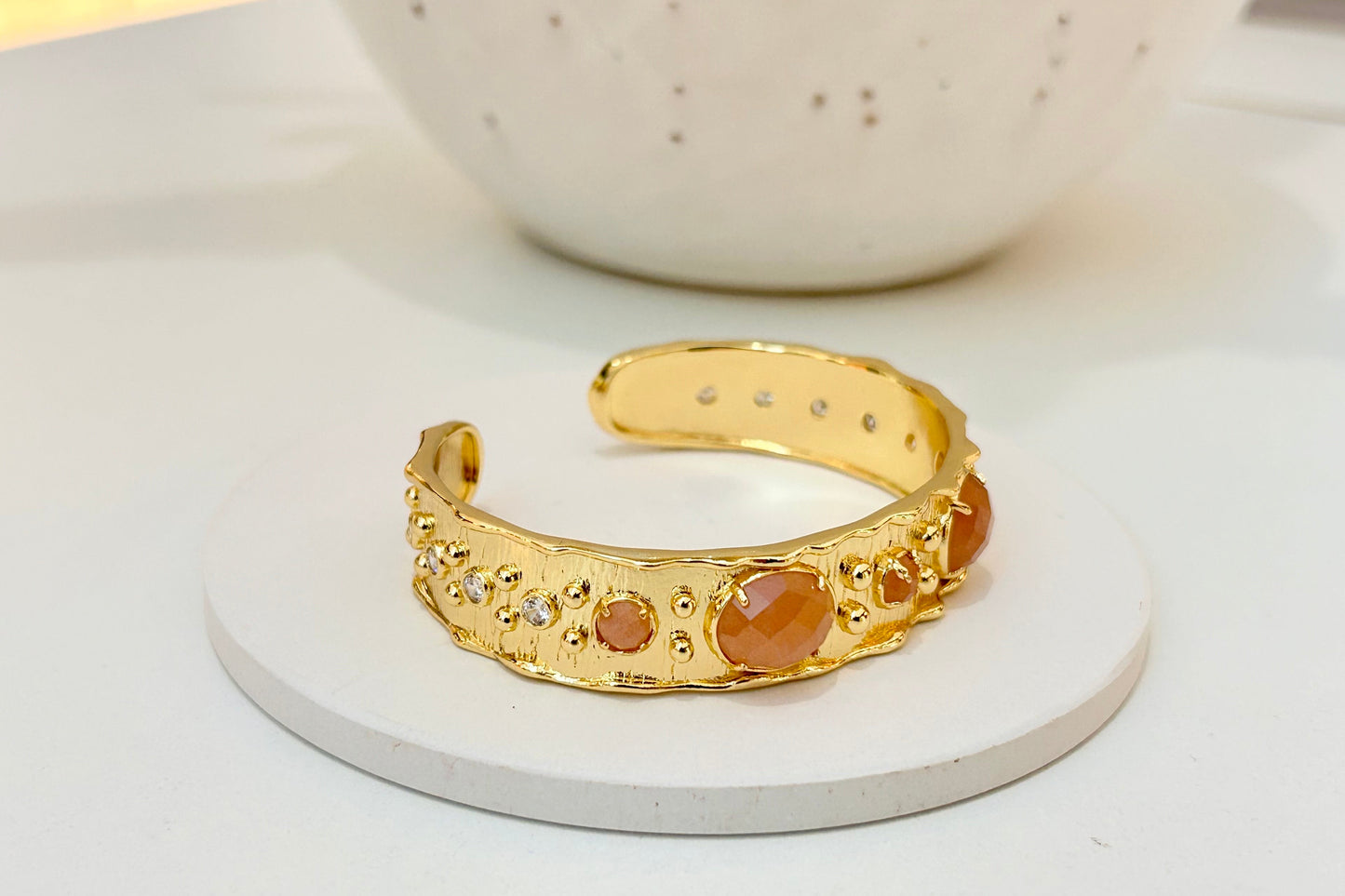 
                  
                    A gold cuff bracelet with textured organic details and round sunstone natural gemstones, displayed on a white circular coaster.
                  
                
