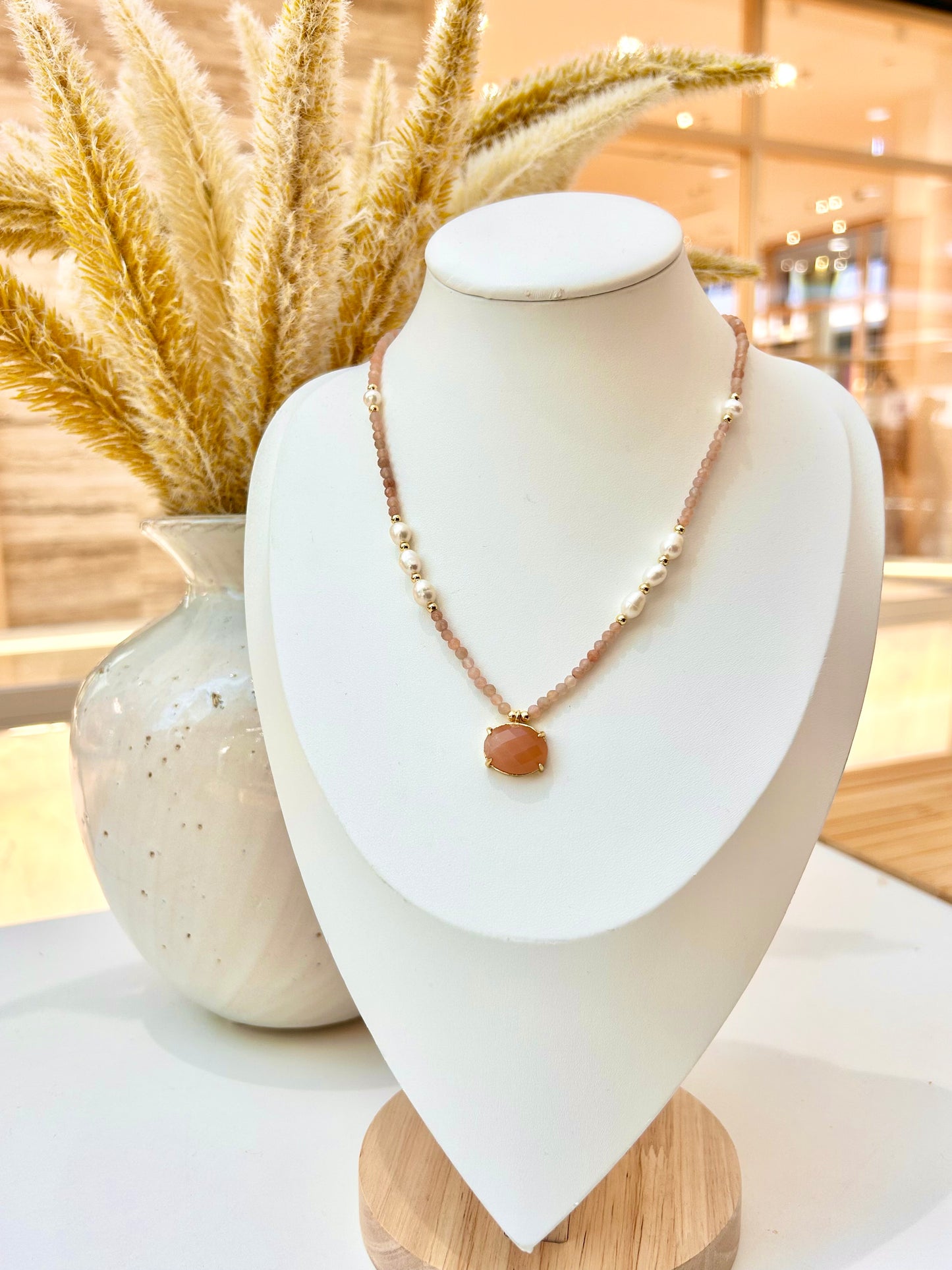 
                  
                    A delicate beaded necklace with a sunstone stone pendant displayed on a white jewelry bust. The necklace features small metallic beads and a textured chain, styled next to a ceramic vase with dried pampas grass in a warm, softly blurred indoor setting.
                  
                