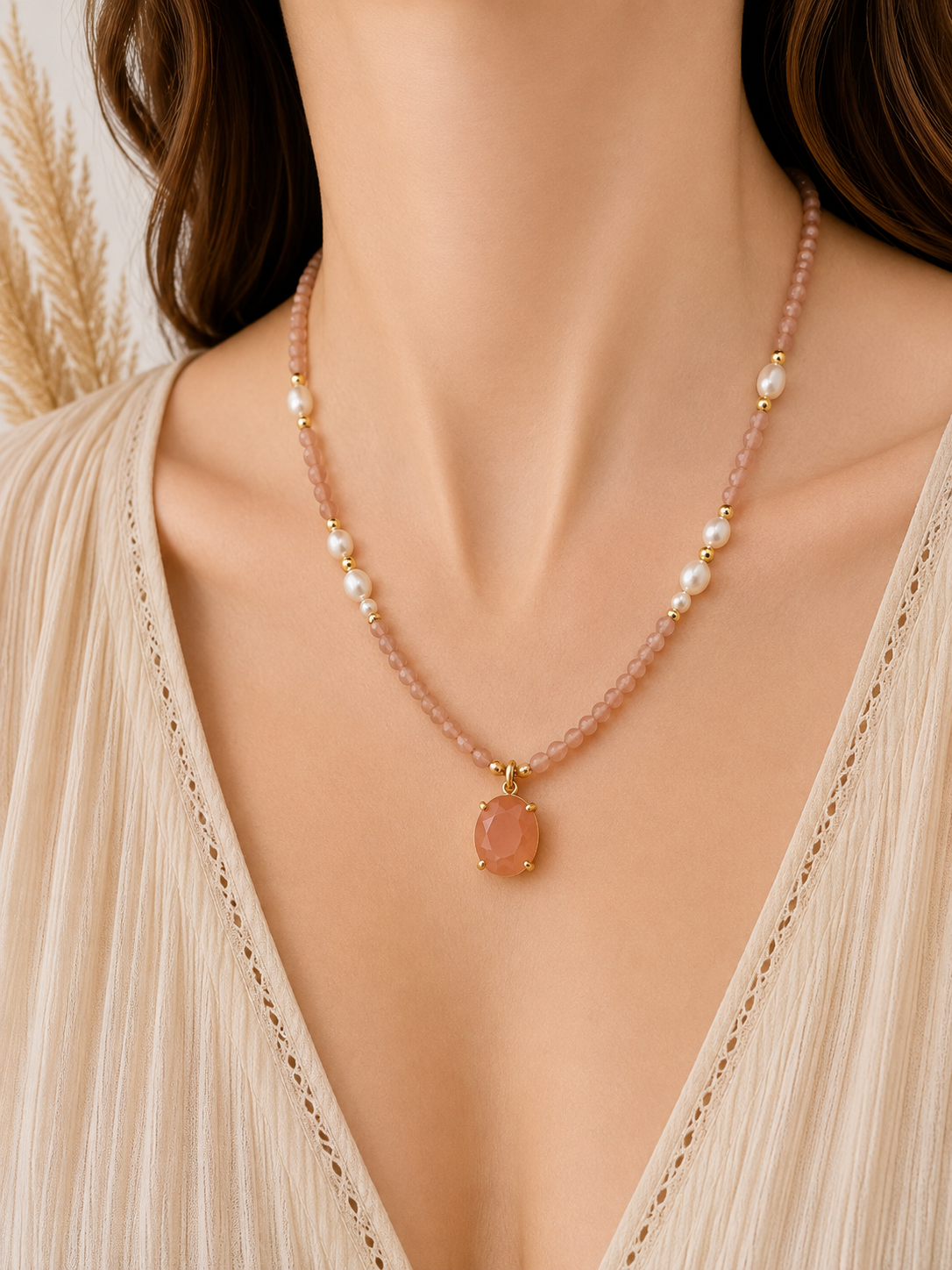 
                  
                    A close-up of a model wearing a delicate sunstone beaded necklace with white freshwater pearl beads and a pink oval pendant in gold-tone settings, paired with a cream-colored pleated top.
                  
                