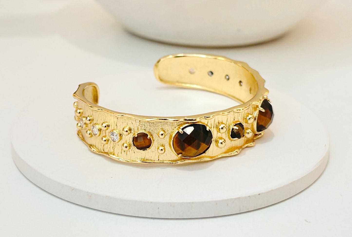 
                  
                    A gold cuff bracelet with large tigers eye  gemstones and small clear rhinestone accents, displayed on a round white coaster with a white speckled bowl in the background.
                  
                