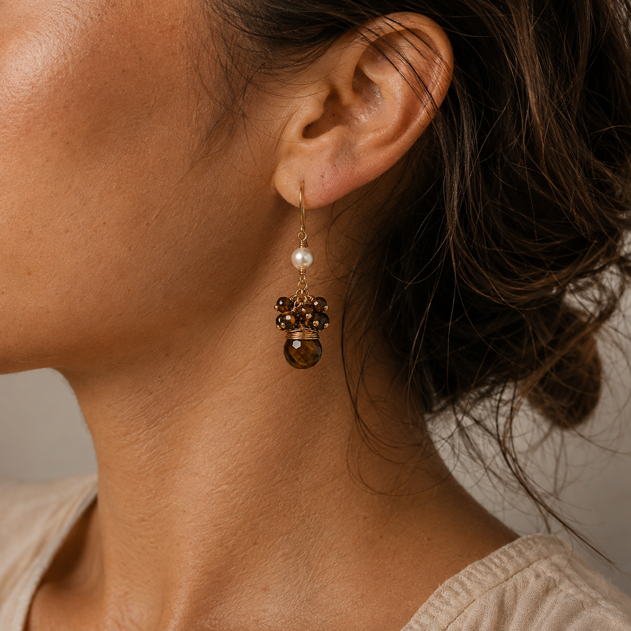 A close-up of a woman's ear and neck wearing a dangling earring. The earring features a gold-tone hook, a small white pearl, and a cluster of dark brown tigers eye  beads with a faceted brown tigers eye gemstone at the bottom.