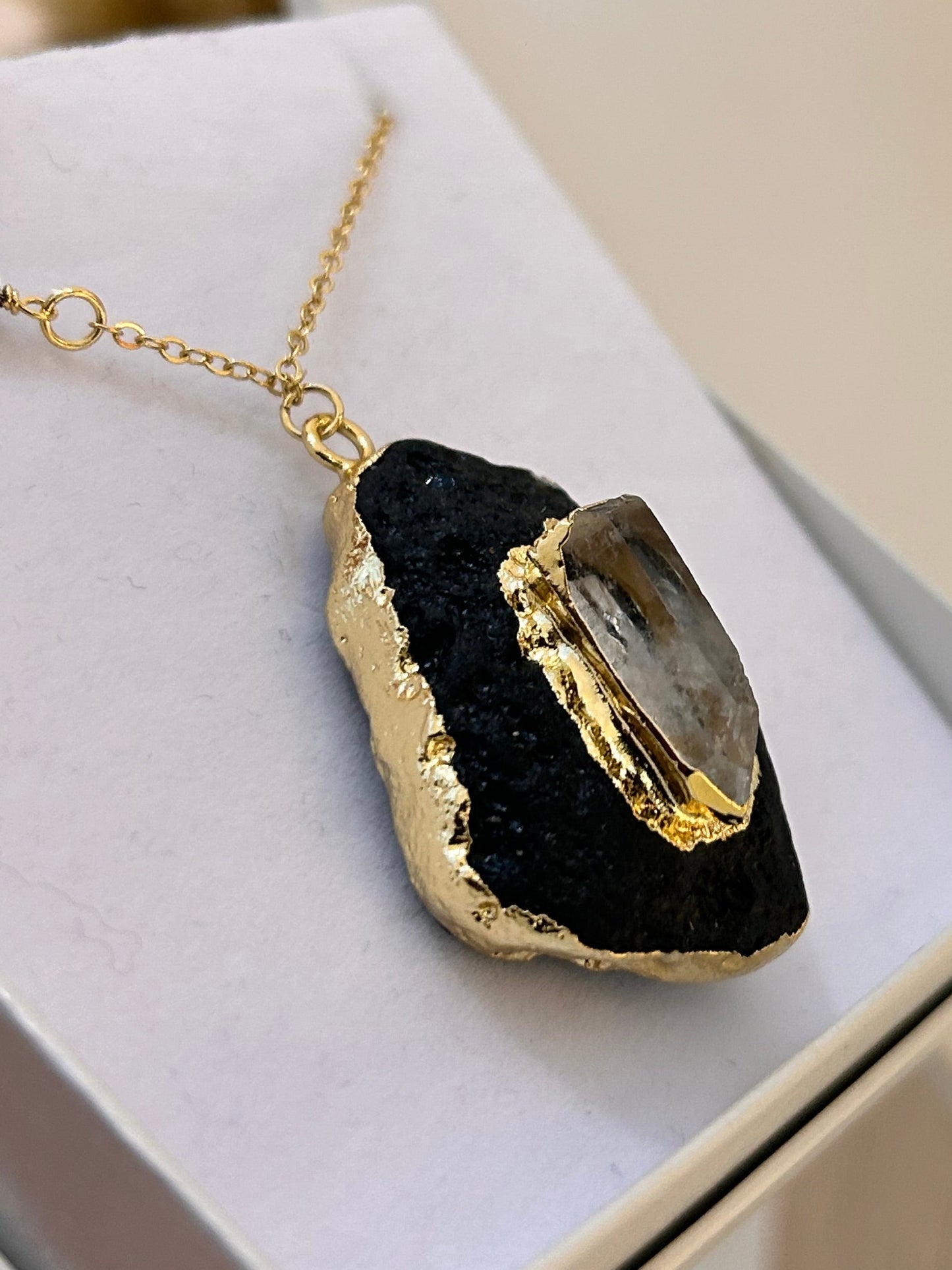 
                  
                    A gold chain necklace with a pendant featuring a black textured top and a polished gold geode-like stone, displayed on a white jewelry box.
                  
                