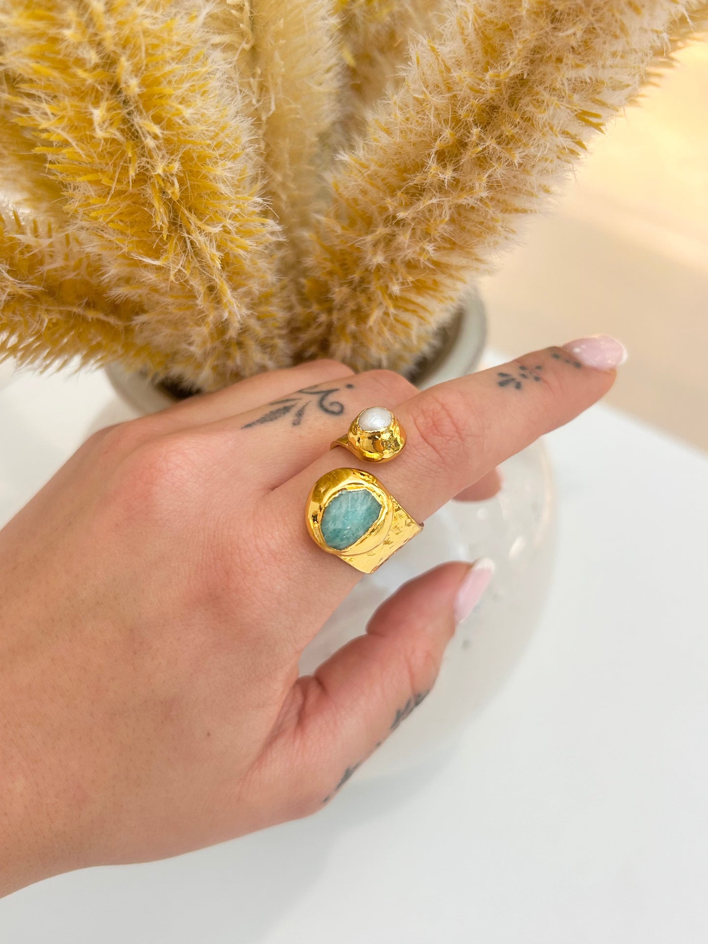 A close-up of a hand wearing two gold rings: one with a round white pearl and the other with a large oval amazonite stone, set against a soft beige background with dried pampas grass.