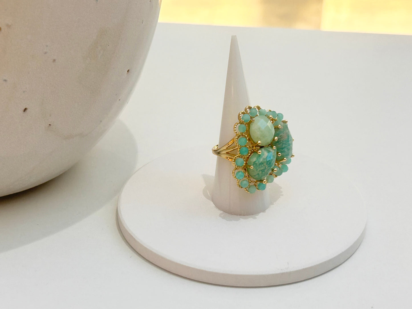 
                  
                    A gold ring with three round light green gemstones and small turquoise-blue accent stones, displayed on a white cone ring holder.
                  
                