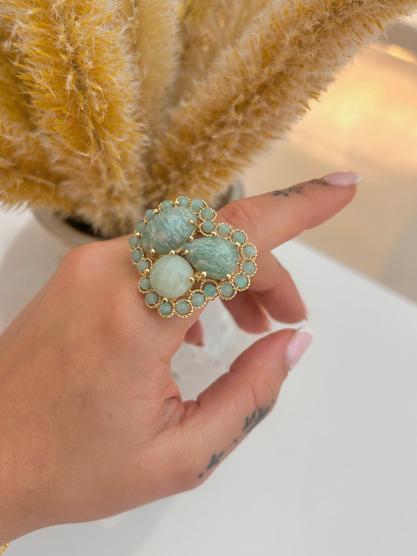 A close-up of a hand wearing a large gold ring with three oval light amazonite stones and a halo of small round green stones, set against a soft beige background with dried grass.