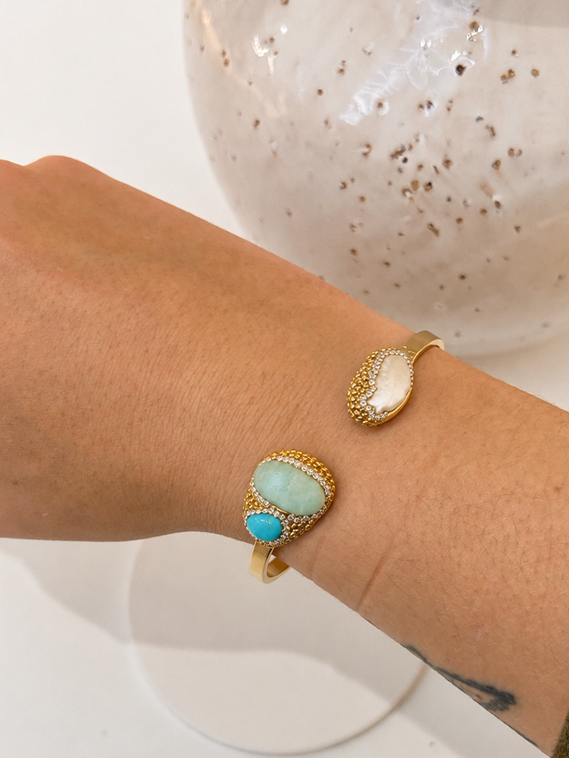 
                  
                    A close-up of a person's wrist wearing gold cuff bangle.  Features a large oval amazonite stone, and the other features a smaller freshwater pearl, both with gold textured borders.
                  
                