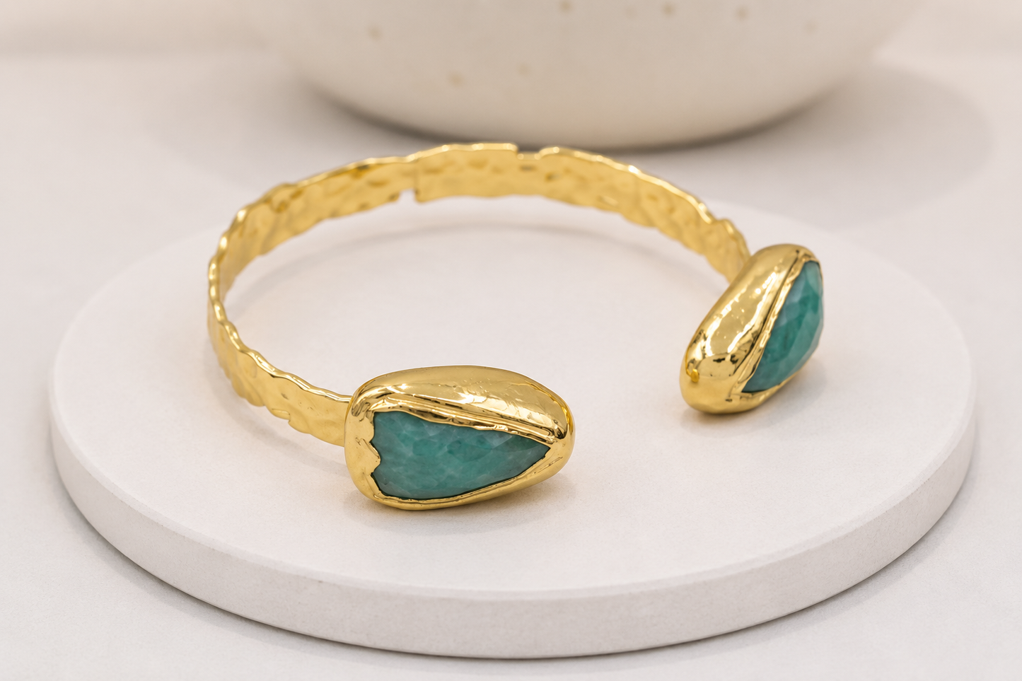 A gold open cuff bracelet with two organic-shaped amazonite stone inlays, displayed on a round white pedestal.