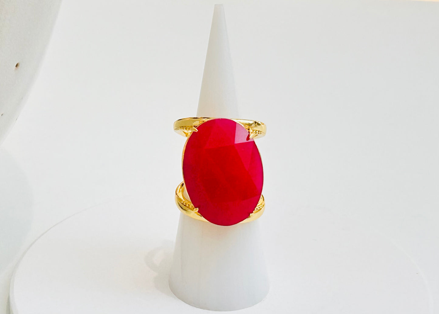 
                  
                    A gold ring with a large oval red gemstone, displayed on a white cone stand against a white background.
                  
                