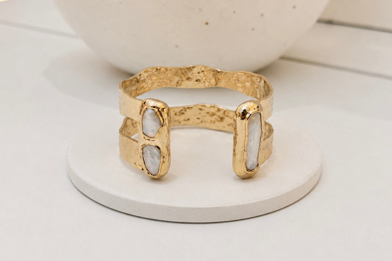 A gold-toned textured cuff bracelet with two vertical oval freshwater pearl inlays, displayed on a round white coaster with a ceramic vase in the background.