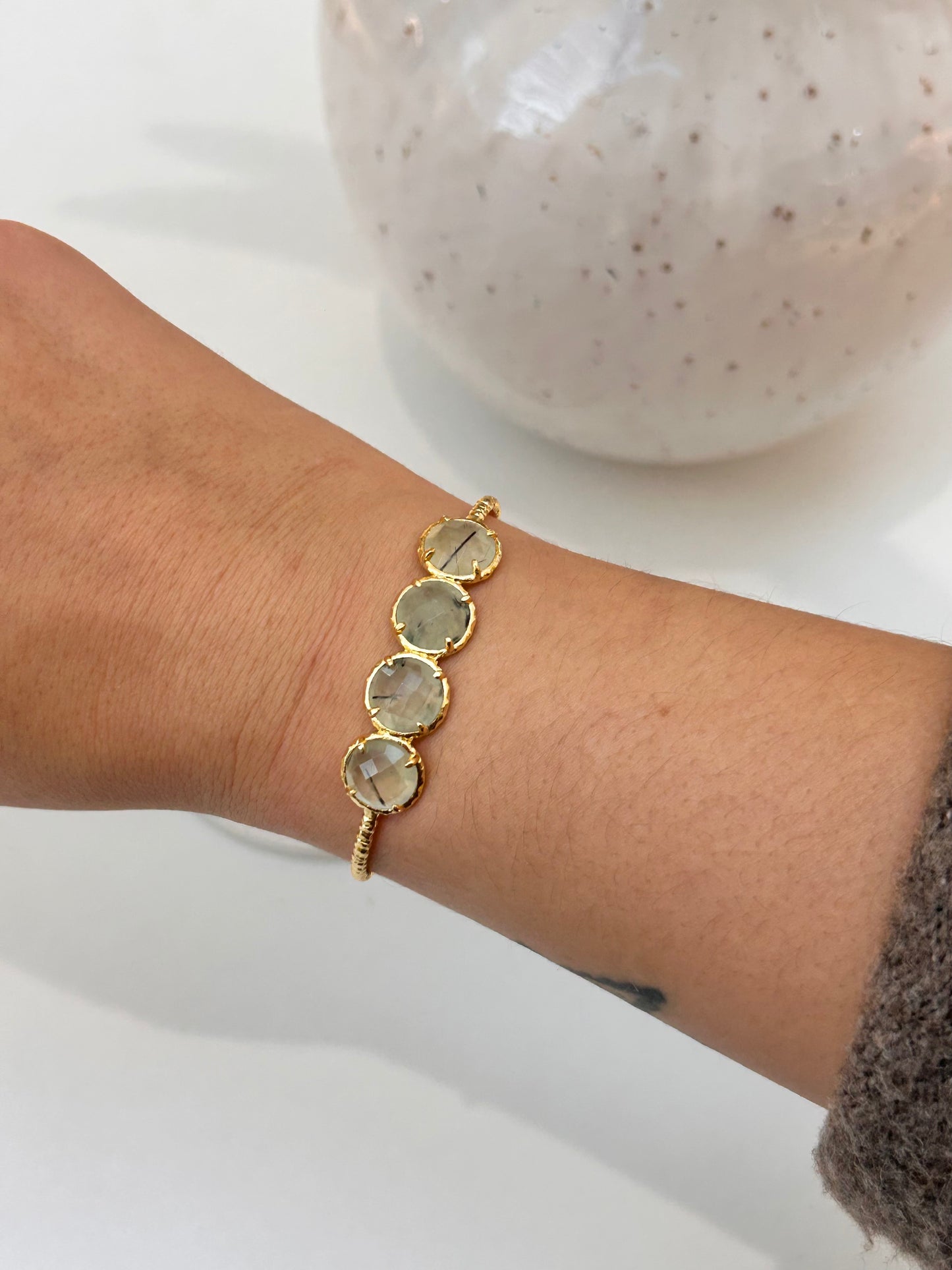 A gold bracelet with four round prehnite stone accents, shown worn on a person's wrist against a white background with a blurred speckled vase.
