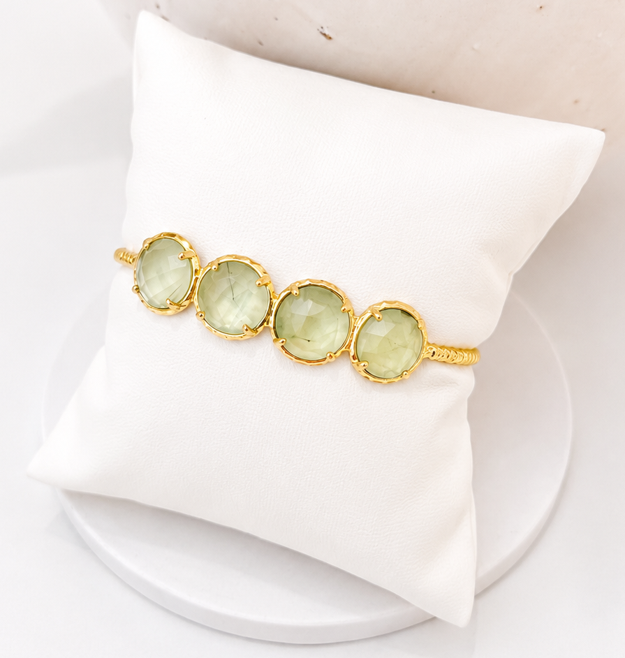 
                  
                    Gold bracelet with prehnite  gemstones on a white cushion against a beige stone background
                  
                