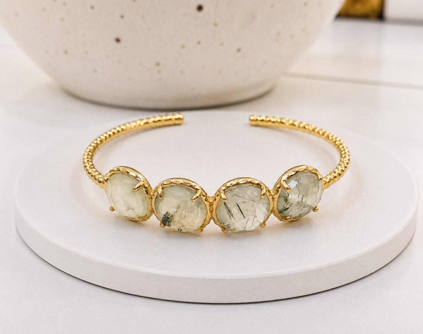A gold-toned open cuff  bracelet featuring four round prehnite crystal displayed on a white round tray with a speckled ceramic bowl in the background.