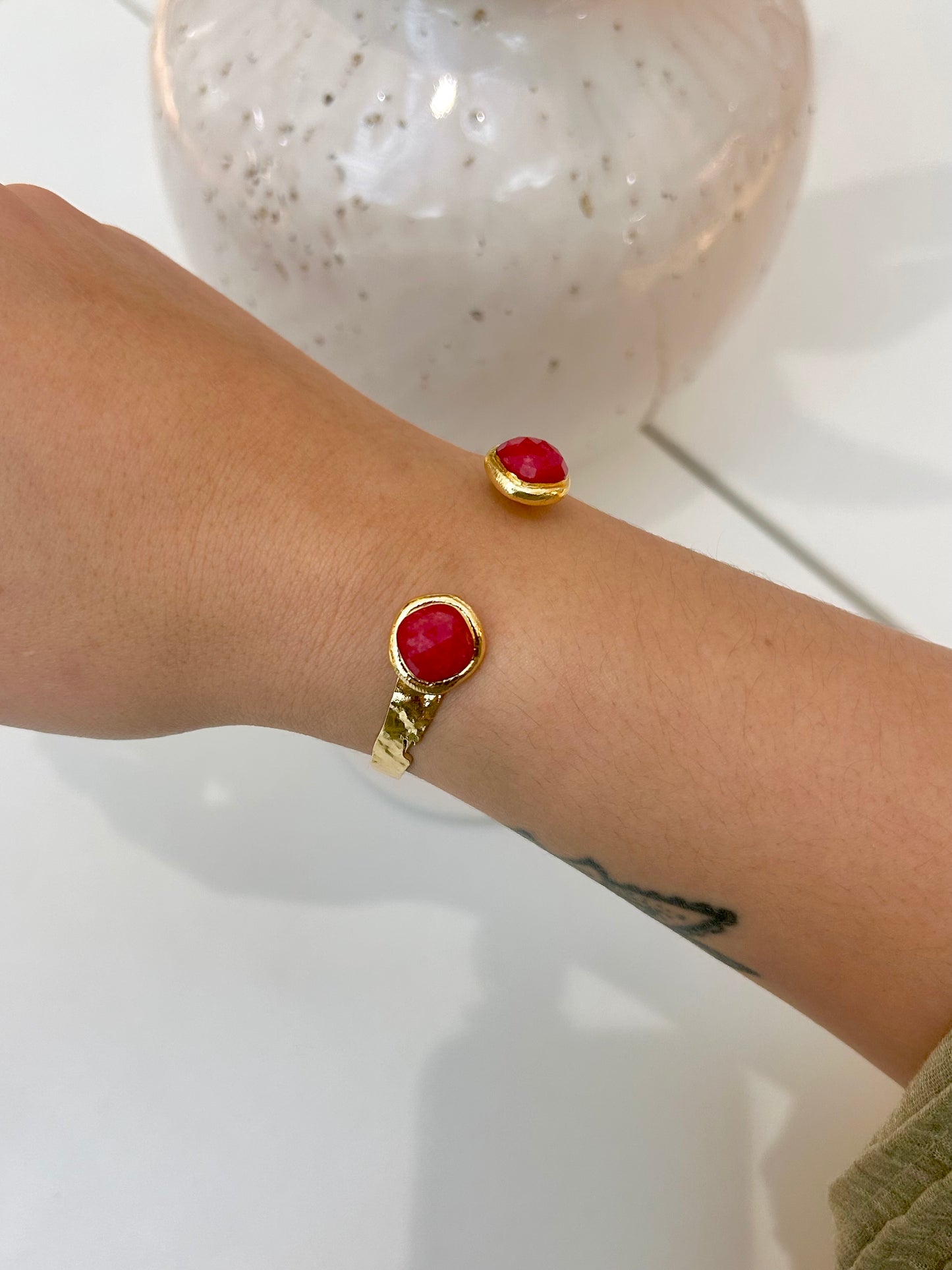 A close-up of a person's wrist wearing two gold-toned bracelets, each featuring a round red jade stone, with a white speckled ceramic vase blurred in the background.
