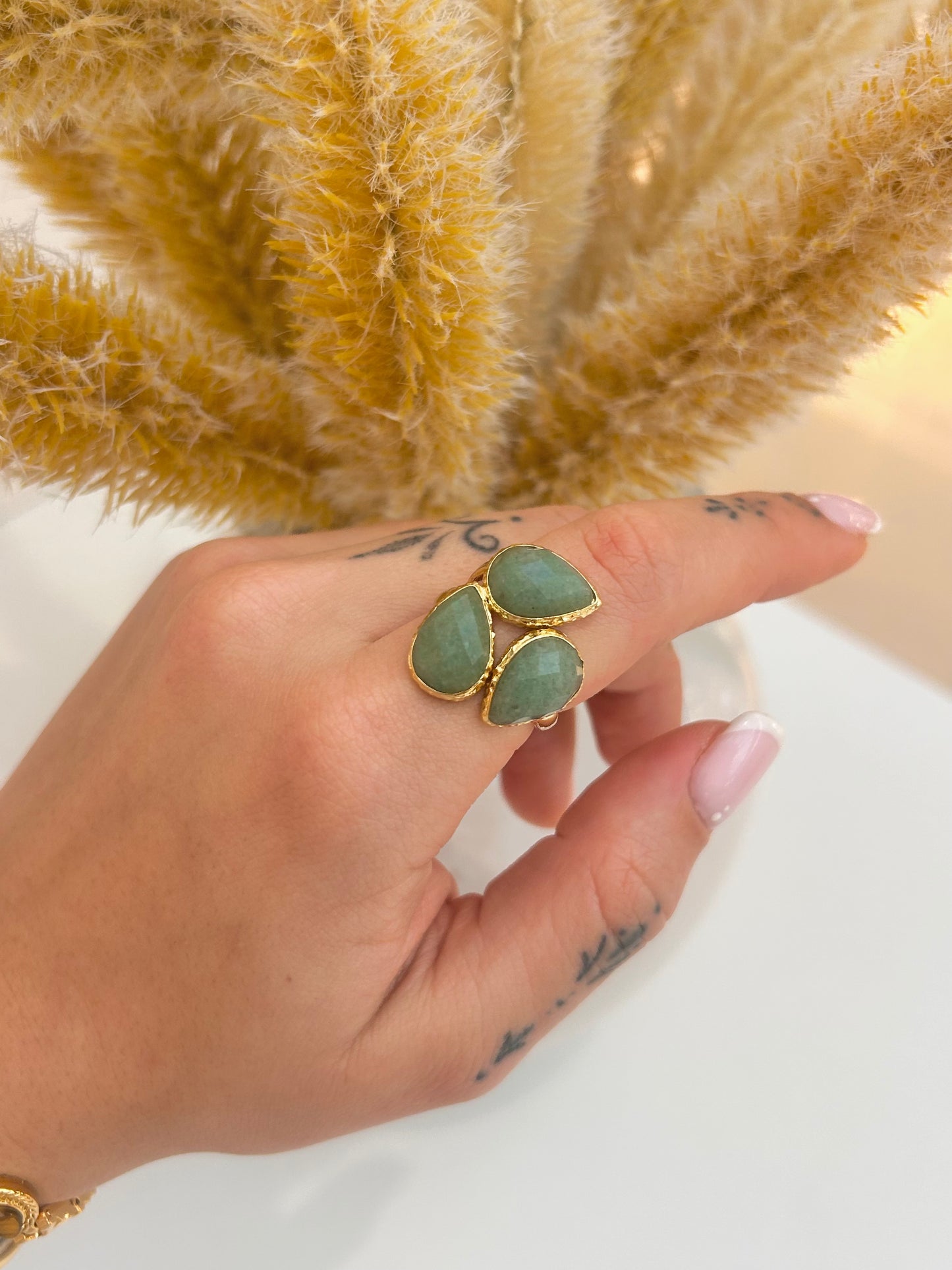 A close-up of a hand wearing a gold ring with three green teardrop-shaped green aventurine stones, set against a background of dried pampas grass.