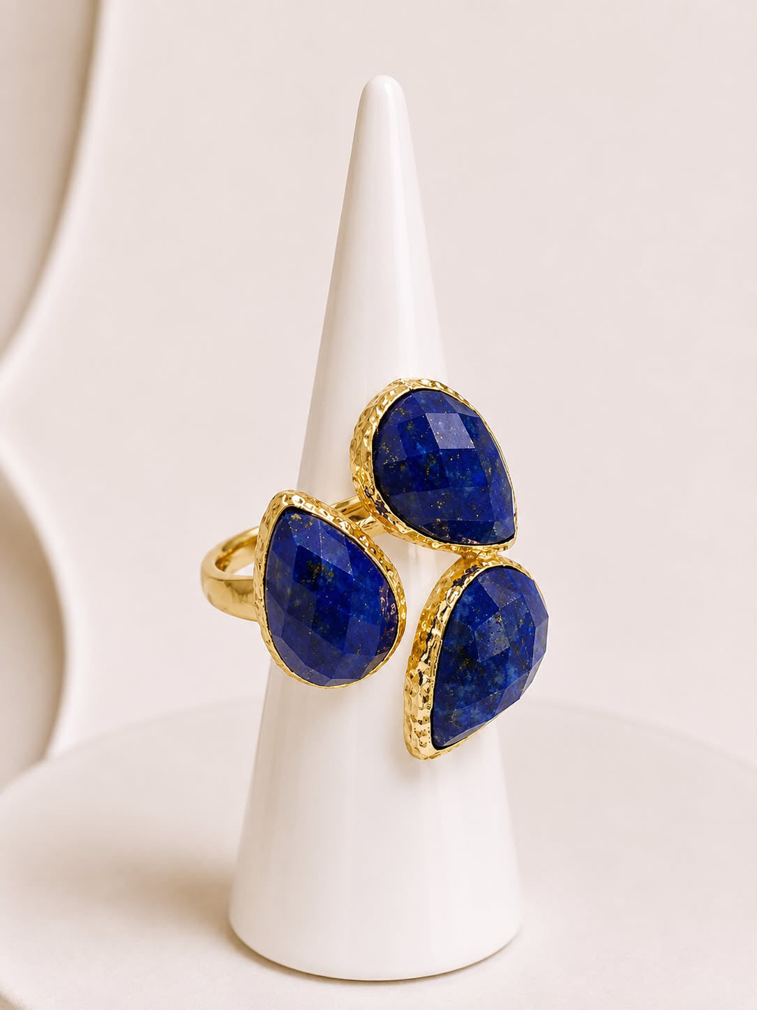 A gold ring with three large teardrop-shaped lapis lazuli gemstones, displayed on a white cone stand against a light beige background.