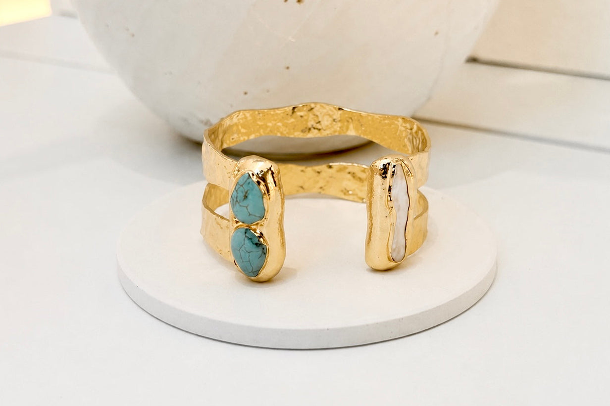 Gold-plated double-band open cuff bracelet featuring two turquoise stones and a long freshwater pearl in organic settings, displayed on a white jewellery stand.
